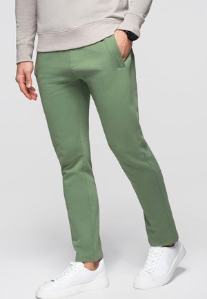 Osoba ubrana w wąskie spodnie w kolorze szałwii, beżową bluzę, białe trampki, z jedną ręką w kieszeni i czarnym zegarkiem na nadgarstku.