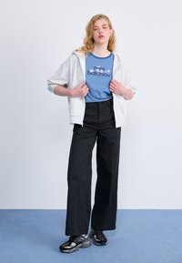 Hoodie gris zippé, t-shirt graphique bleu avec "new balance", pantalon large noir et baskets noires avec des accents gris.