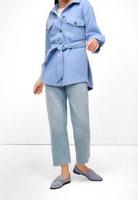 Světlomě modrá košilová bunda s páskem v pase a ramenními nášivkami, doplněná o světlé džíny s rovnými nohavicemi a modré mokasíny se zlatými akcenty.