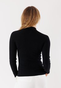 Donna con lunghi capelli castano chiaro, indossa un top aderente nero a costine a maniche lunghe e pantaloni bianchi, girata di spalle contro uno sfondo semplice.