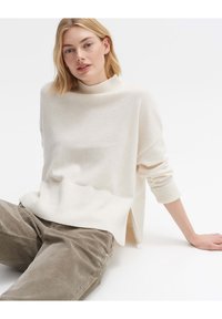 Hellbeige Pullover mit hohem Kragen, gerippten Bündchen und seitlichem Schlitz, kombiniert mit olivgrünen Hosen. Weiche Textur und lockere Passform.