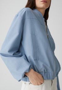 Veste bomber bleu clair avec une texture lisse, fermeture éclair et taille élastique. Présente des manches surdimensionnées et des poches latérales.
