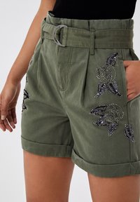 Olivgrüne Shorts mit Gürtel, hochgekrempelten Säumen und schwarzer, perlenbestickter Blumenmuster, getragen mit einem schwarzen Oberteil, Hand in der Tasche ruhend.