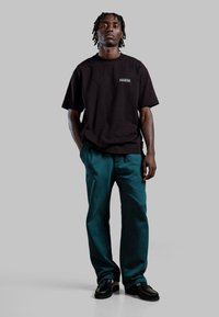 Czarna bawełniana koszulka z małym logo na piersi; w zestawie z turkusowymi spodniami i czarnymi butami. Model stoi na szarym tle.