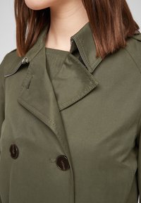 Donna che indossa un trench coat doppiopetto verde oliva con bottoni scuri e capelli castani dritti fino alle spalle, visibile dalla gola al petto.