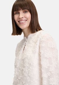 Weißes Blusenshirt mit strukturierten floralen Applikationen, rundem Ausschnitt, Knopfdetails und langen Ärmeln. Das Material scheint leicht und transparent zu sein.