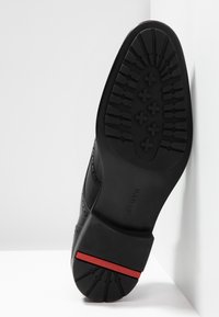Chaussure en cuir noir avec une semelle texturée, présentant un motif sur les côtés et un accent rouge sur le talon.