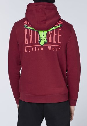 Takakuva henkilöstä, joka pukeutunut viininpunaiseen huppariin, jossa vihreä sukeltajahahmo ja vaaleanpunainen teksti "Chillsee Active Wear" selässä.