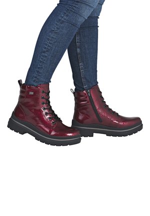 Glänzende dunkelrote Schnür-Stiefeletten mit seitlichen Reißverschlüssen, getragen zu eng anliegenden blauen Jeans vor weißem Hintergrund.