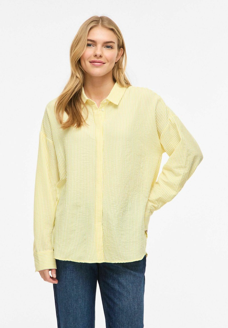 Jeune femme aux cheveux blonds portant une chemise ample à rayures jaune pâle boutonnée et un jean bleu, debout avec une main sur la hanche.