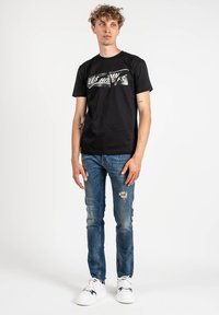 Camiseta de algodón negra con texto metálico "LES HOMMES" en el frente, combinada con jeans de mezclilla desgastados en azul claro y zapatillas blancas.