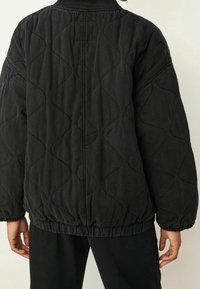 Czarna pikowana kurtka z falistym wzorem, elastycznym dołem i ściągaczem. Materiał wydaje się teksturowany i trwały z luźnym krojem.