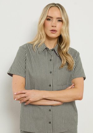 Blondynka o falowanych włosach, ubrana w szarą, krótkorękawową koszulę w białe pionowe paski, stojąca z założonymi ramionami na jednolitym tle.