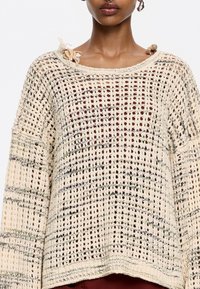Beiger Strickpullover mit offener Maschenware und einem Gittermuster. Auffällige Details sind ein dekorativer Blumenakzent am Halsausschnitt.