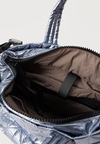 Καπιτονέ μεταλλική ασημί τσάντα με ανοιχτό φερμουάρ που αποκαλύπτει μπεζ εσωτερικό ύφασμα και πολλαπλές θήκες, συμπεριλαμβανομένης θήκης με φερμουάρ και ελαστικής λουρίδας.