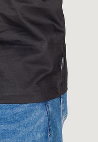 T-shirt in cotone nero con taglio dritto, etichetta nella cucitura laterale e cuciture visibili. Abbinato a jeans in denim blu. Design semplice.