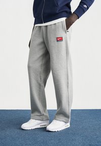 Γκρι φόρμα με πλαϊνές τσέπες, διακοσμημένη με μπλε και κόκκινο λογότυπο Nike. Συνδυασμένη με λευκά αθλητικά παπούτσια σε φόντο μπλε χαλιού.