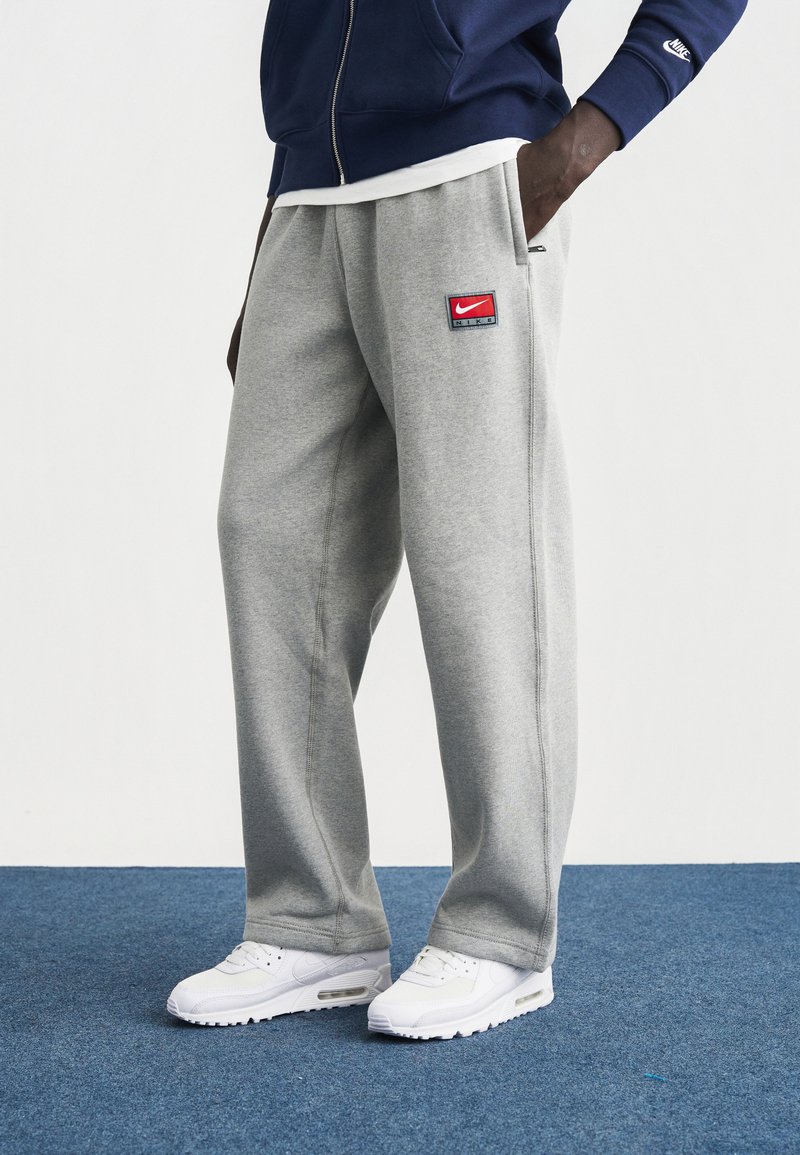 Γκρι φόρμα με πλαϊνές τσέπες, διακοσμημένη με μπλε και κόκκινο λογότυπο Nike. Συνδυασμένη με λευκά αθλητικά παπούτσια σε φόντο μπλε χαλιού.