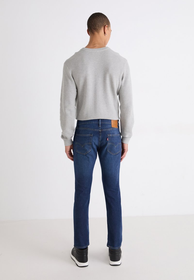 Maglione grigio lavorato a maglia, jeans in denim blu e sneakers nere. Texture a coste sul maglione, etichetta in pelle sui jeans, design dal taglio standard.