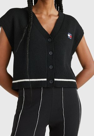 Vrouw die een zwart, mouwloos gebreid vest met knopen draagt, met witte strepen en een klein Tommy Jeans-logo op de borst, gecombineerd met een zwarte broek.