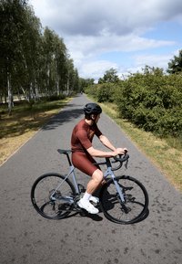 Racefiets met een lichtblauw frame, zwarte wielen en aflopende sturen. Fietsrijder in een rode fietskleding, witte sokken en een zwarte helm, op een pad.
