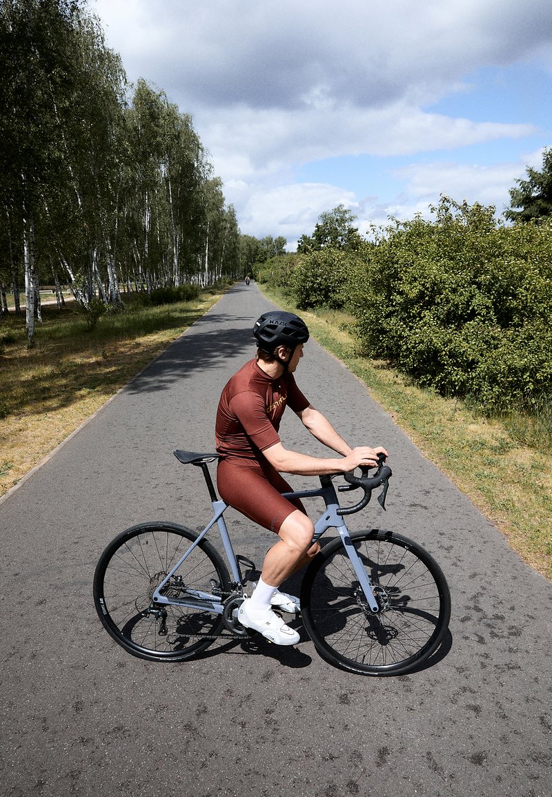 Racefiets met een lichtblauw frame, zwarte wielen en aflopende sturen. Fietsrijder in een rode fietskleding, witte sokken en een zwarte helm, op een pad.
