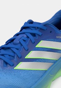 Μπλε αθλητικό παπούτσι με επάνω μέρος από δίχτυ, υφή στην επιφάνεια, ασημένιες ρίγες και πράσινες λεπτομέρειες στη μεσαία σόλα. Διαθέτει επίπεδα κορδόνια και ενισχυμένη φτέρνα.