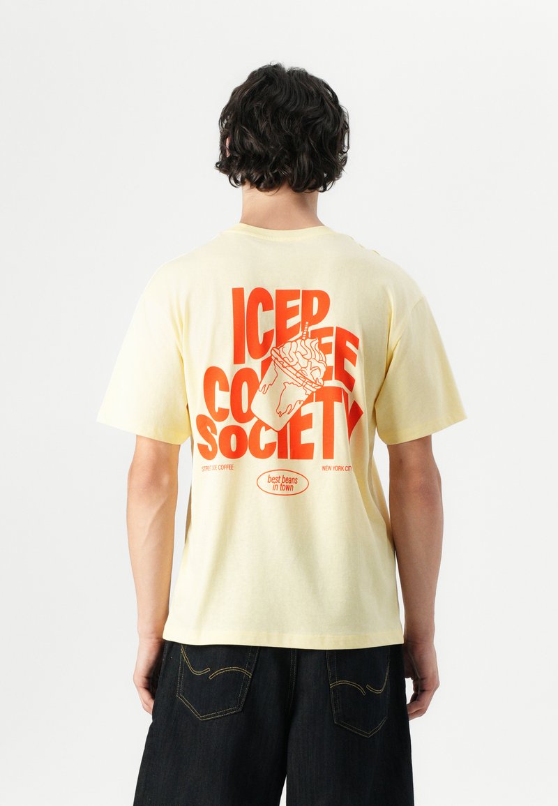 Dzeltena kokvilnas T-kreklu ar sarkano grafisko dizainu mugurpusē, kas ietver tekstu "ICED COFFEE SOCIETY" un dzēriena ilustrāciju.