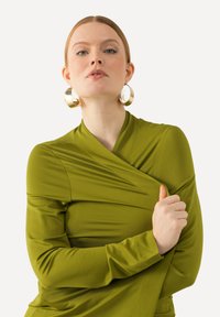 Langärmlige, wickelartige Bluse in Olivgrün, mit einer glatten Textur und seitlichen Drapierungen. Große, runde goldene Creolen runden das Design ab.