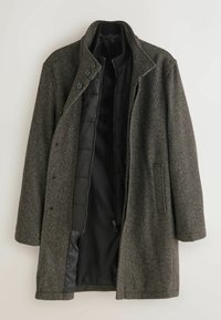 Lång mörkgrå ullrock med svart quiltad innerfoder, knappar och dragkedja, samt två sidofickor, visas öppen.