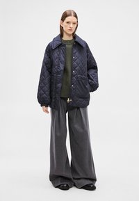 Καπιτονέ μπλε ναυτικό μπουφάν με μεγάλο γιακά, κουμπιά με κλειδώματα και μια μπροστινή τσέπη. Συνδυασμένο με πράσινο λαδί πουλόβερ και άνετο γκρι παντελόνι.