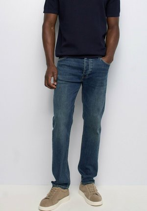 Hombre con vaqueros azules, camisa de manga corta azul marino oscuro y zapatillas beige, de pie con una mano en el bolsillo sobre fondo blanco.