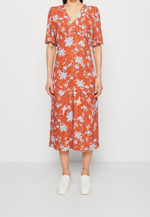 Kvinde iført en rust-orange midikjole med blomsterprint og knapper foran samt hvide slip-on sko, stående foran en ensfarvet baggrund.