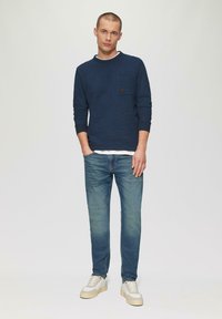Marineblaues Sweatshirt mit Struktur, Rundhalsausschnitt und Brusttasche, kombiniert mit hellblauen Jeans und weißen Sneakers mit beigen Akzenten.