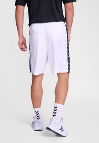 Weiße Sportshorts mit schwarzen Chevron-Streifen an der Seite, kombiniert mit weißen Sneakers und gemusterten Socken. Der Stoff wirkt leicht.