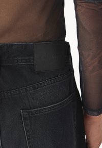Black denim jeans with a textured finish, featuring a stitched pocket and a branded leather patch on the waistband. Sheer black mesh top visible.