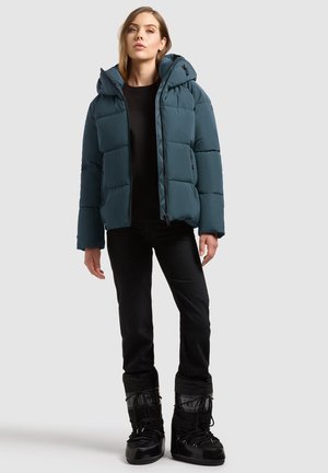 Femme portant une doudoune turquoise avec capuche, pantalon noir, bottes noires et haut noir, debout devant un fond clair uni.