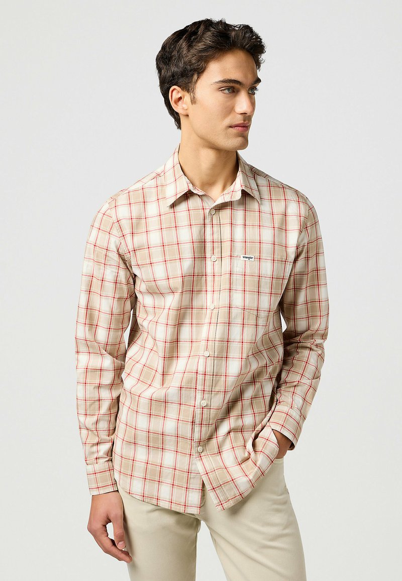 Camisa de cuadros beige con detalles rojos, de manga larga, con un bolsillo en el pecho y un pequeño logo de la marca en el bolsillo.