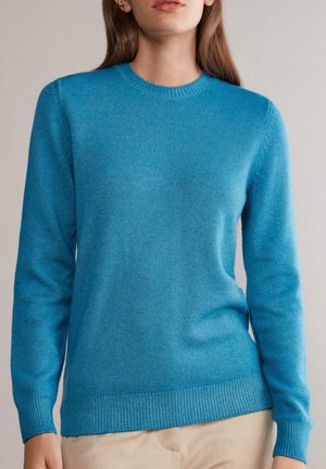 Donna che indossa un maglione a maniche lunghe di colore blu brillante con girocollo e polsini e orlo a coste, abbinato a pantaloni beige.