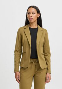 Olivově zelený měkký látkový blazer s těsným střihem, se zářezovými klopami a dvěma předními kapsami, kombinovaný s černým topem a odpovídajícími tepláky.