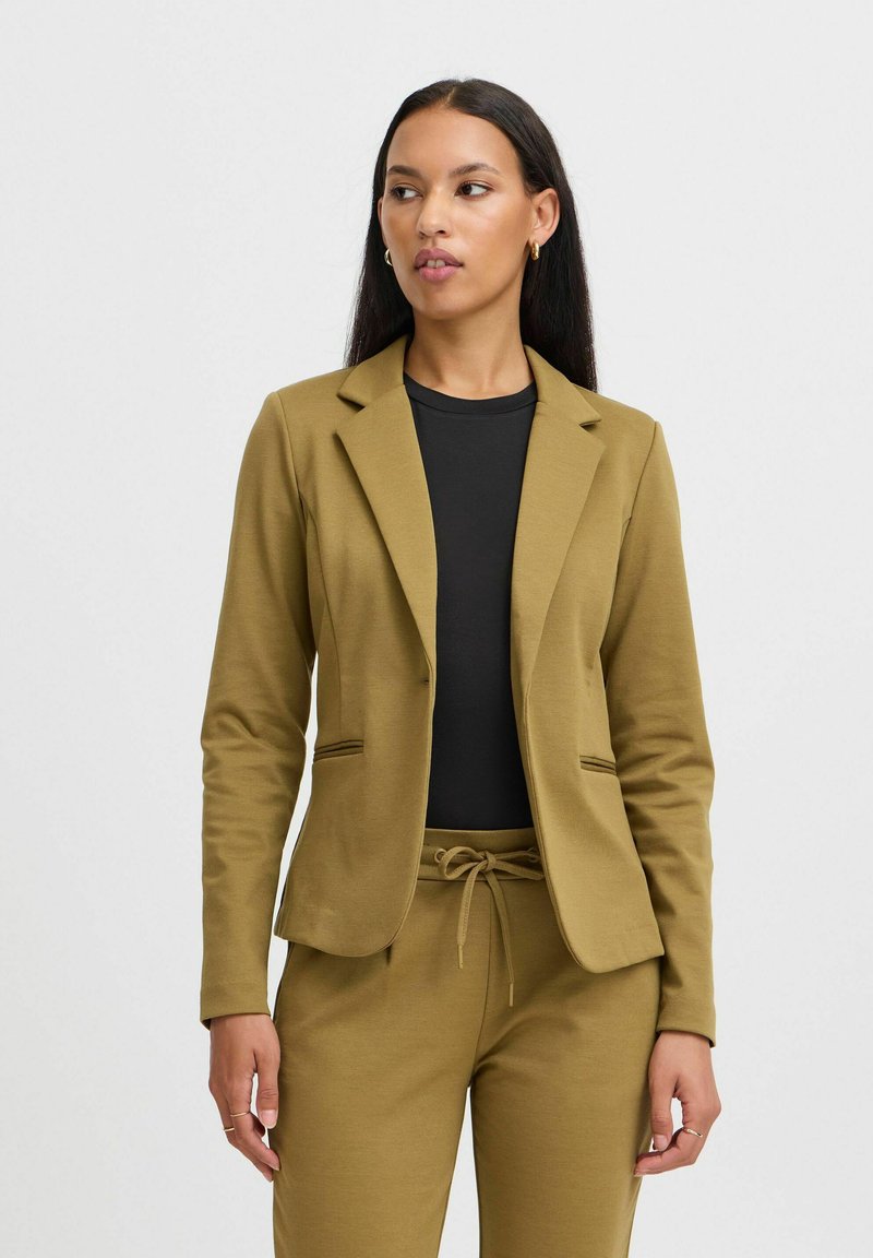 Vrouw draagt mosterdgele blazer en bijpassende broek over een zwarte top, staand tegen een effen lichte achtergrond, kijkt opzij.