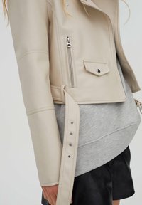 Beige Leder-Shortjacke mit seitlichem Reißverschluss und Druckknopfverschluss. Sie hat eine taillierte Passform und eine glatte Textur, über einem grauen Sweatshirt getragen.