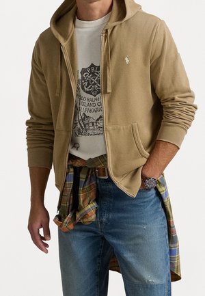 Man wearing beige zip-up hoodie over white graphic t-shirt with plaid shirt tied around waist and blue jeans, hand in pocket.