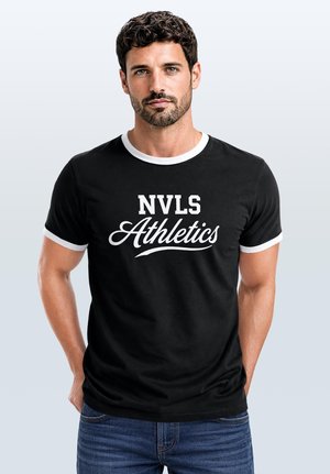 Man with short curly dark hair and beard wearing black and white ringer T-shirt with "NVLS Athletics" text standing against plain light background.