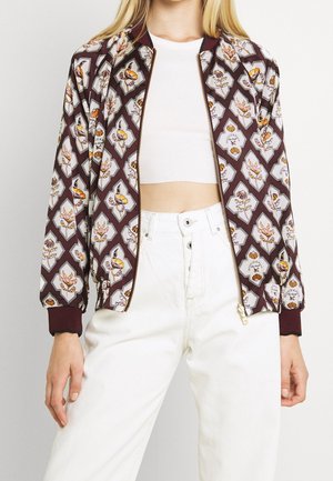 Personne portant une veste bomber bordeaux à motifs floraux, un top court blanc et un pantalon blanc taille haute, montrée du cou aux hanches.