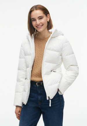Mujer sonriente con cabello corto y castaño, lleva una chaqueta acolchada blanca, suéter de punto marrón, vaqueros oscuros y cinturón negro sobre fondo blanco.