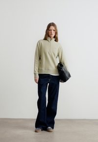 Νεαρή γυναίκα στέκεται μπροστά σε απλή τοίχο φορώντας μπεζ φούτερ με φερμουάρ, φαρδιά σκούρα τζιν, γκρι παπούτσια και κρατώντας μια μαύρη δερμάτινη τσάντα κάτω από το μπράτσο της.