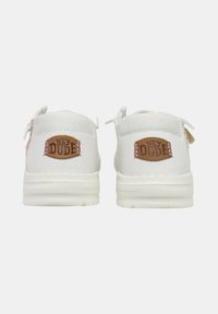 Witte canvas sneakers hebben een ronde neus, een gestructureerd oppervlak en een bruine leren logo patch aan de achterkant met "HEY DUDE" in reliëf.