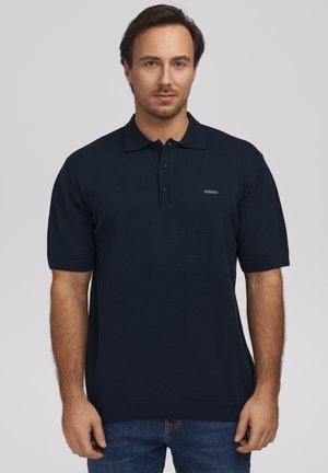 Mężczyzna z krótkimi brązowymi włosami i brodą, ubrany w granatową koszulkę polo z krótkim rękawem i niebieskie dżinsy na jednolitym szarym tle.