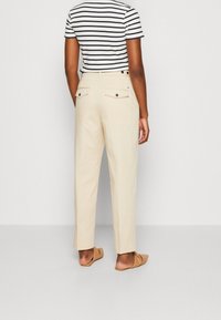 Pantalones beige a medida con dos bolsillos traseros y detalles de botones, combinados con una camisa a rayas y sandalias marrones tipo slide.